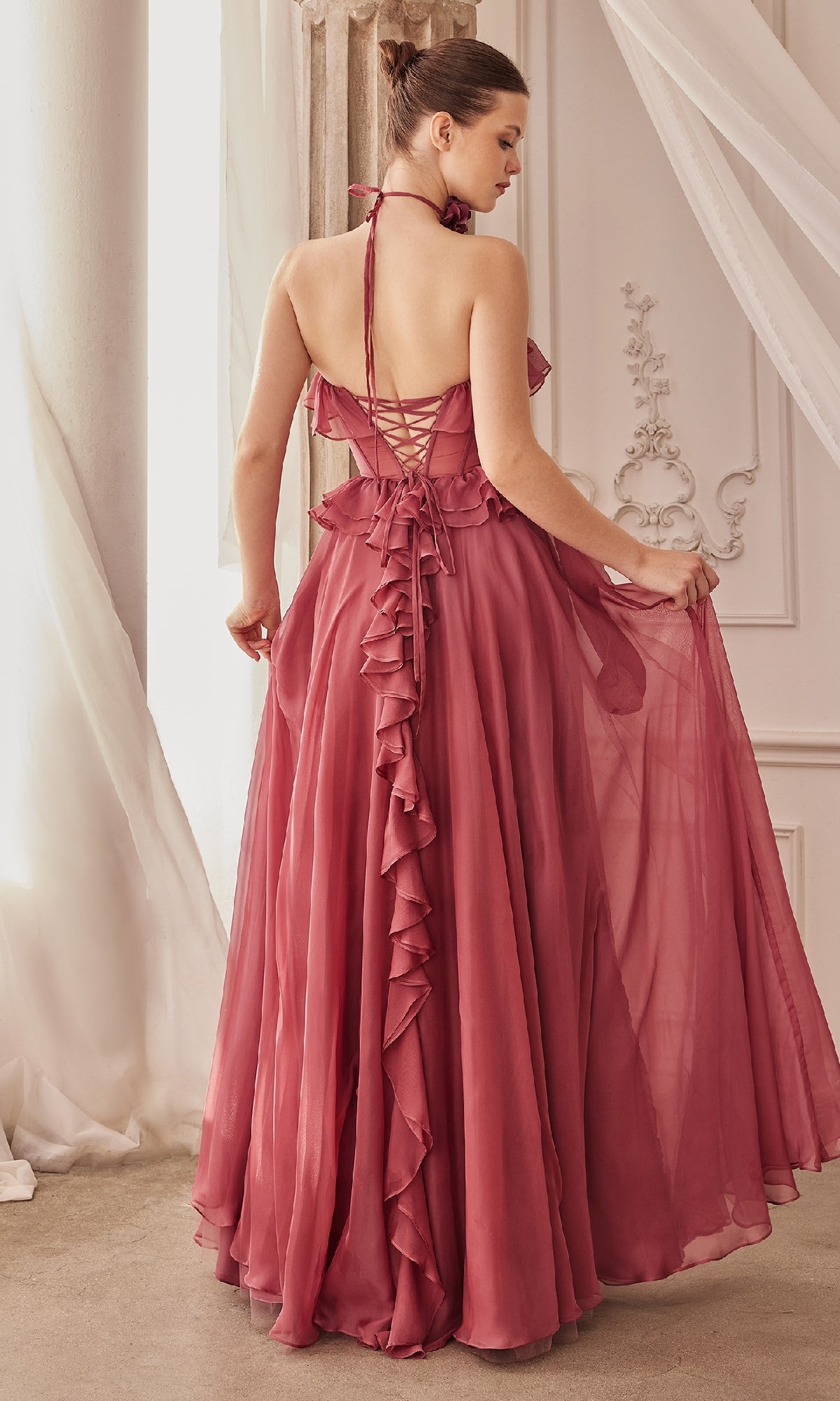 A woman stands with her back to the camera in an elegant room, wearing the Andrea and Leo A1341 Strapless Ruffled Prom Dress—a floor-length rose gown featuring an open back, ruffle details, and a lace-up back.